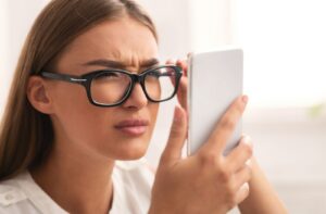A young woman wearing eyeglasses is squinting while looking at her cell phone screen