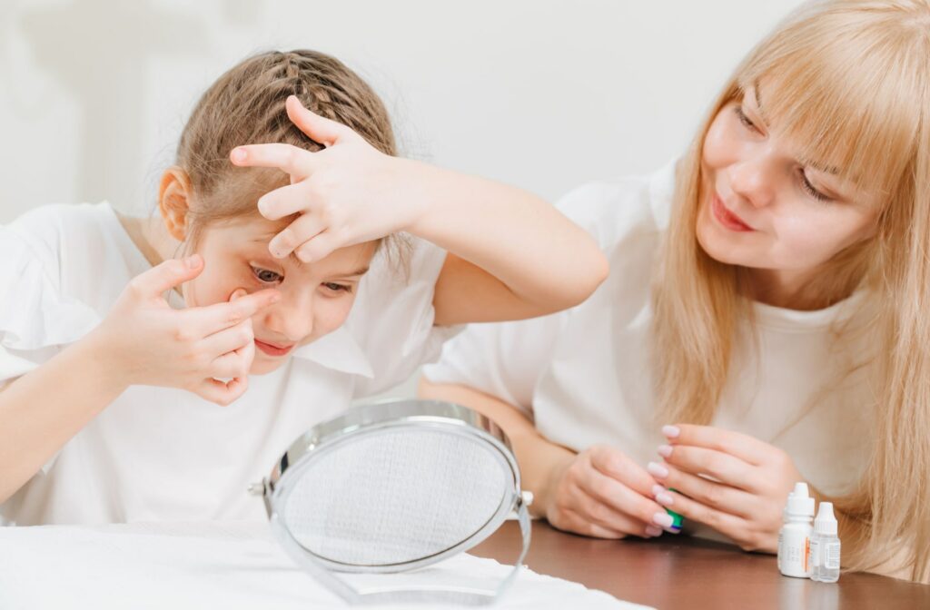 A young girl putting a contact lens on her eye in front of a small mirror while being supervised by a woman beside her.