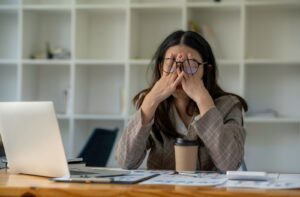 A woman sitting at her desk with her laptop open. She's rubbing her eyes underneath her glasses due to eye pain and headaches.