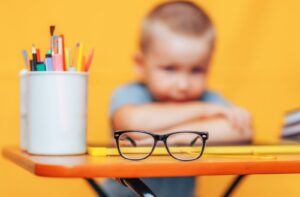 A child sits with his hands crossed over his desk staring at his glasses, which are in focus, in front of him.
