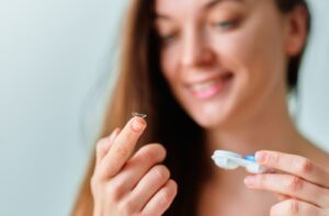 A woman holding a contact lens case in her left hand while balancing a contact lens on her right index finger.