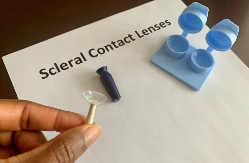 A person holds a lens inserter while a suction cup and lens case sit on a paper reading "scleral contact lenses"