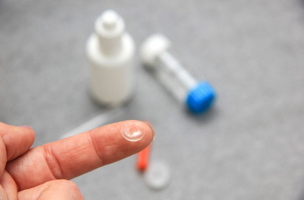 A person holds a contact lens on their fingertip. The contact lens case and a bottle of cleaning solution sit on a table in the background.