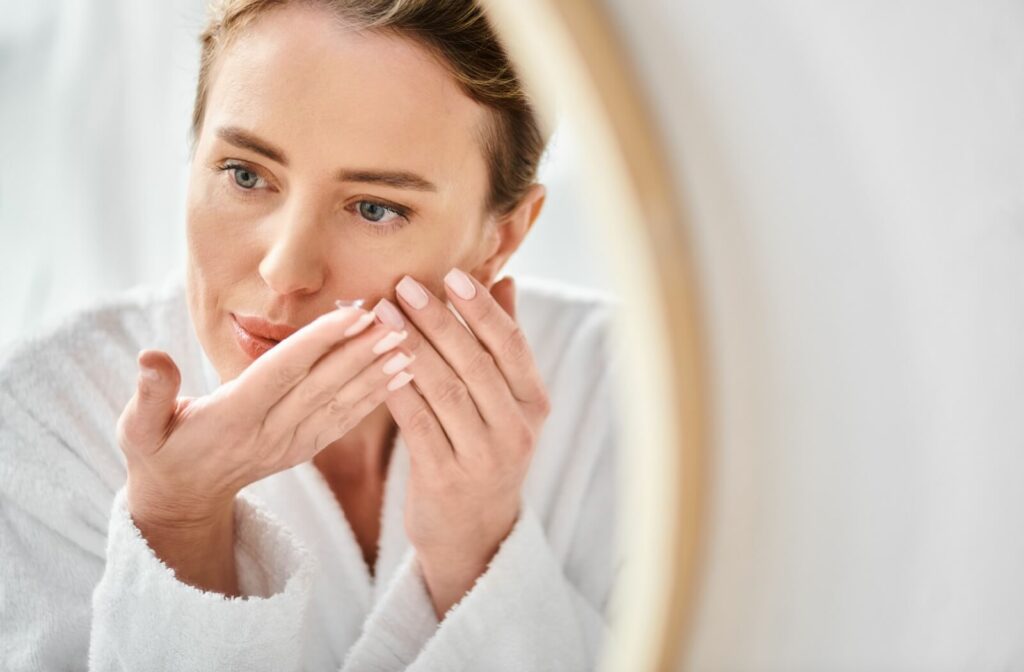 A person in a bathrobe stands in front of a bedroom mirror, inserting a contact lens.
