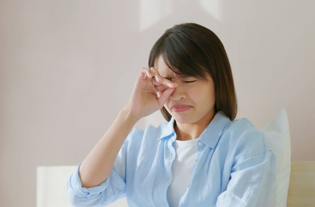A woman sitting in bed in the morning, rubbing her irritated eyes, possibly experiencing symptoms of dry eye discomfort after waking up.