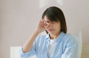 A woman sitting in bed in the morning, rubbing her irritated eyes, possibly experiencing symptoms of dry eye discomfort after waking up.