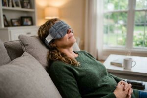 Adult reclining on a sofa wearing a gray quilted eye mask, relaxed, in a living room with a steaming mug nearby.