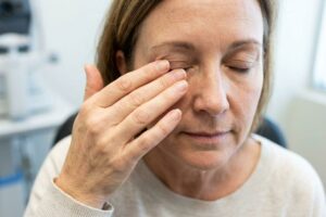 Middle-aged adult rubbing a closed eye with one hand while seated in a clinic, eyes shut.