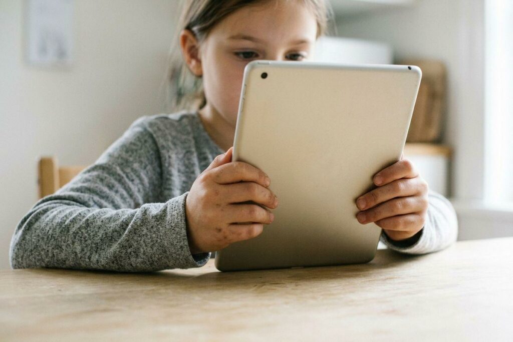 Child sitting at a wooden table, holding and looking intently at a tablet device indoors.